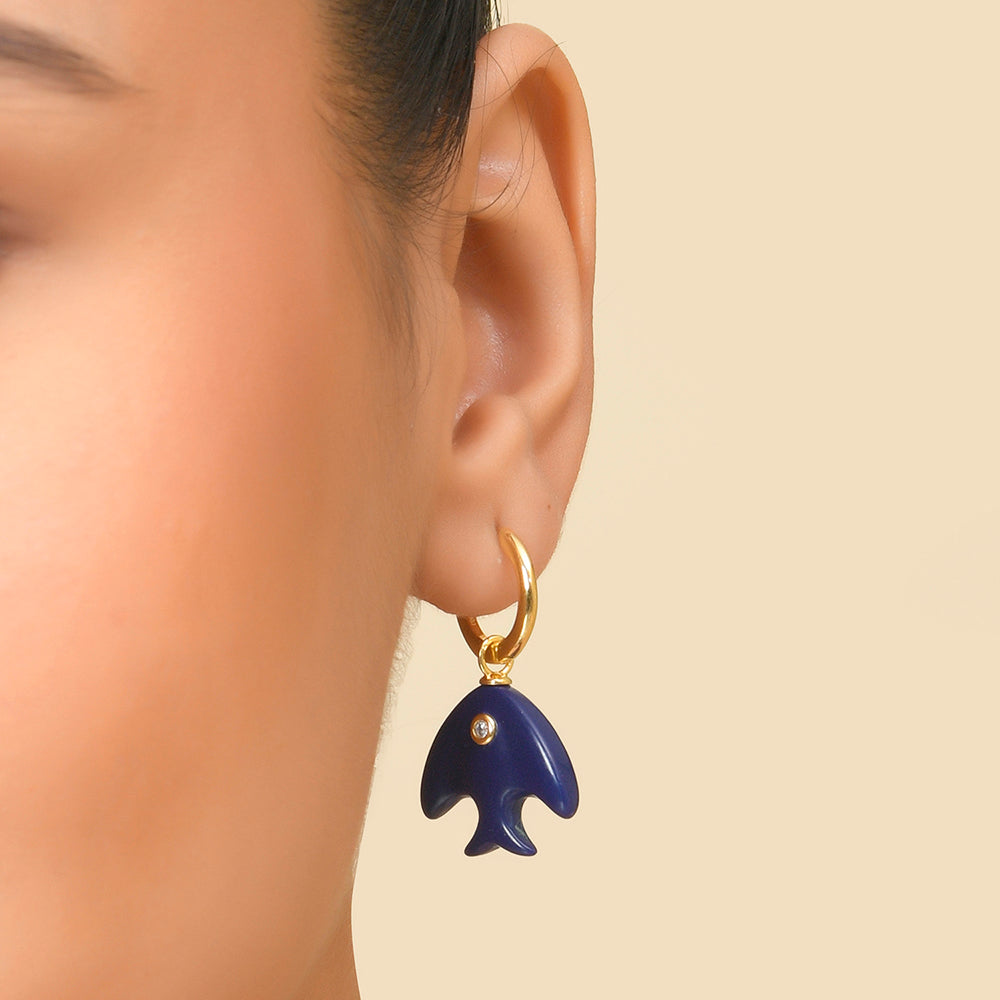 A person wearing a pair of deep blue earrings shaped like a fish, with gold-colored hooks.