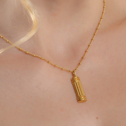 A gold-colored pillar-shaped necklace on a person's neck, showcasing a small, ornate charm.
