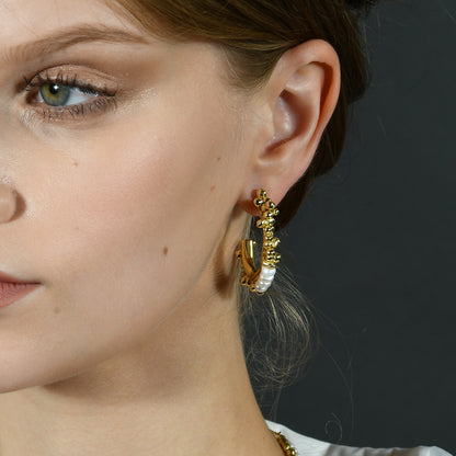 A close-up image of a woman wearing hoop earrings with natural freshwater pearls and golden-hued metal pellets.