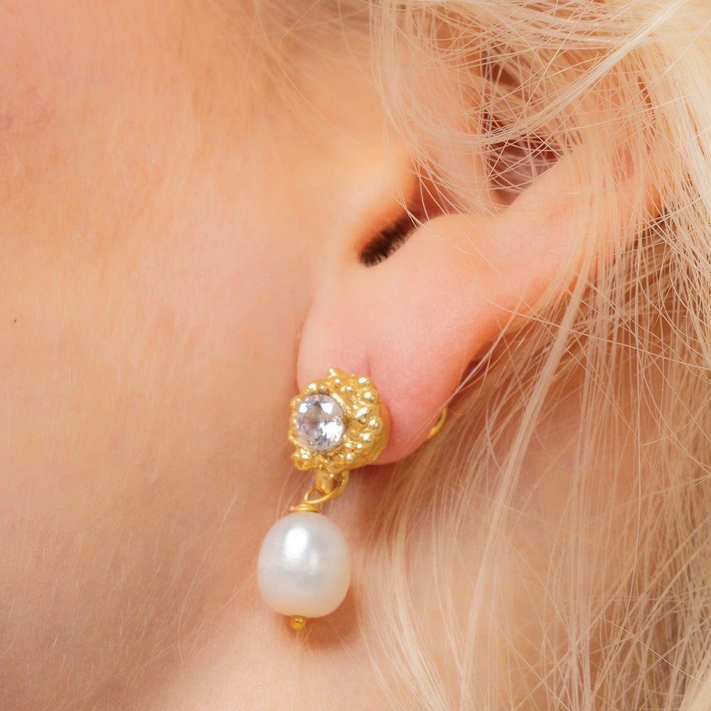 A close-up image of a woman's ear wearing a gold-colored stud earring with a lustrous pearl drop By Arvino