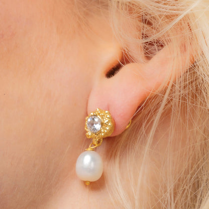 A close-up image of a woman's ear wearing a gold-colored stud earring with a lustrous pearl drop By Arvino