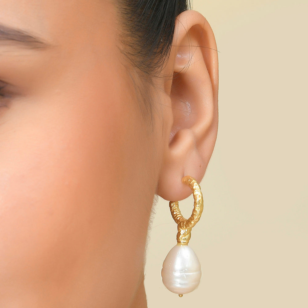 A close-up image of a woman's ear wearing a gold-colored drop hoop earring with a white pearl.