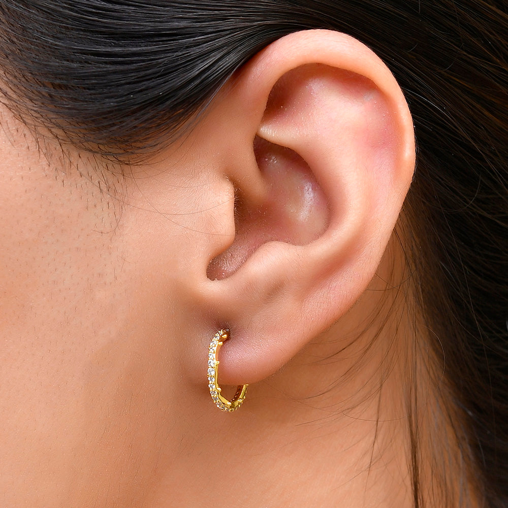 A close-up image of a woman's ear wearing a gold-colored hoop earring studded with cubic zirconia stones.